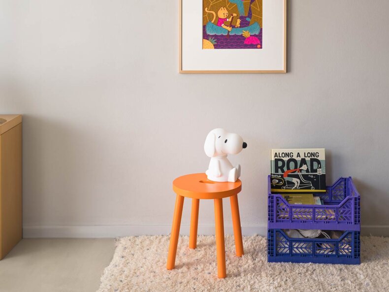 Ein heller Raum mit einem orangefarbenen Kinderhocker KIHO von filipok, auf dem eine Snoopy-Lampe von MR MARIA sitzt. Der Hocker steht auf einem hellen, flauschigen Teppich. Rechts daneben befinden sich zwei lilafarbene, gestapelte Kunststoffkisten AY KASA, die Kinderbücher enthalten, darunter ein Buch mit dem Titel 'Along a Long Road'. An der Wand darüber hängt ein buntes Poster mit dem Titel "Cat in CANOE ", Siebdruck von Nana Rausch, mit einem Motiv von einem Kätzchen in einem Boot.